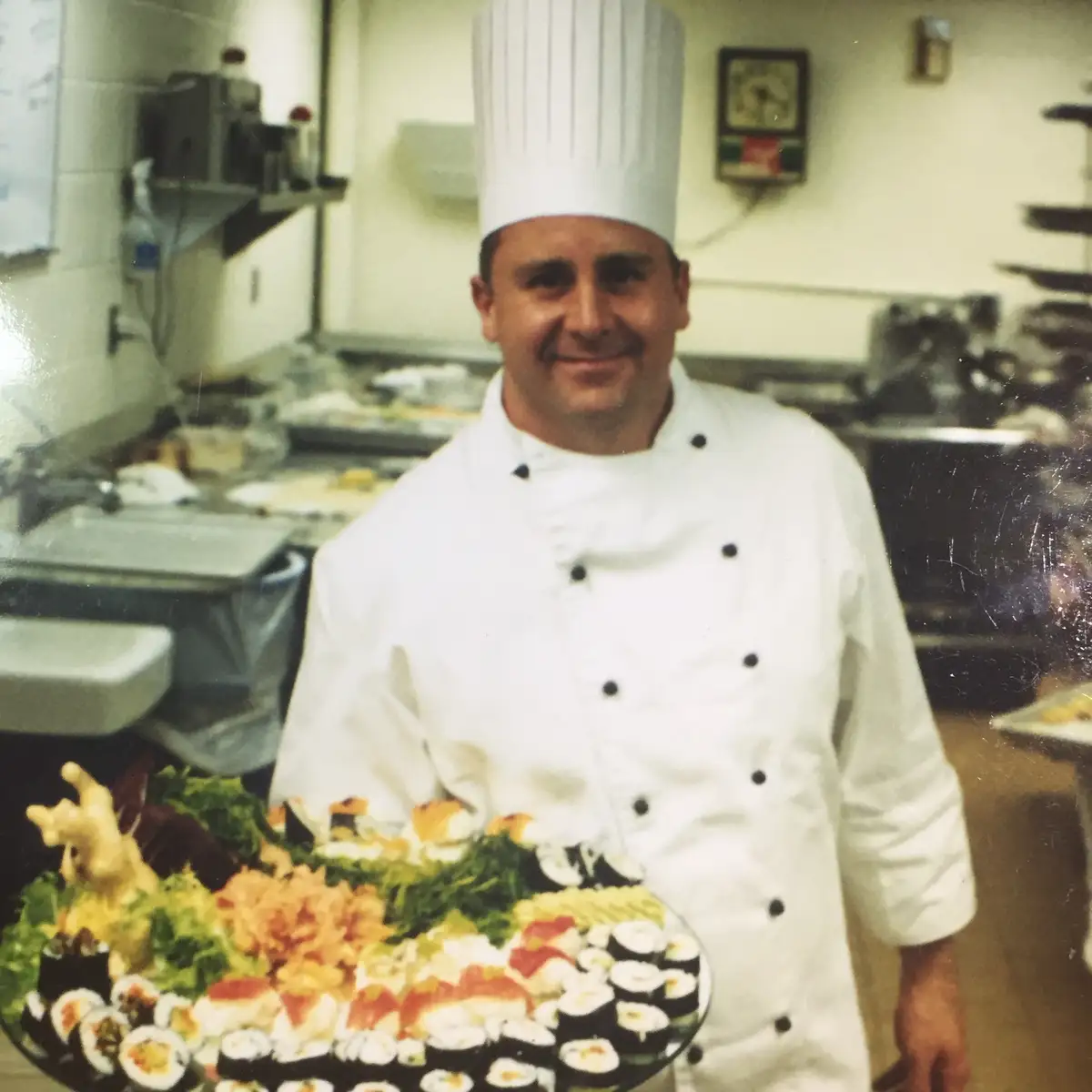 A man in a white chef's uniform and tall hat stands in a kitchen holding a large platter of sushi, garnished with lettuce…