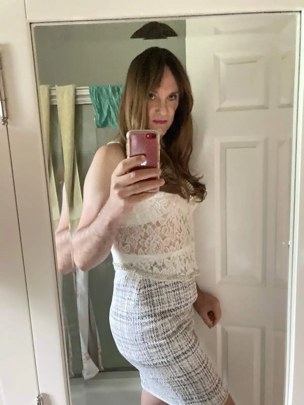 A person with long brown hair and pink lipstick takes a mirror selfie in a bathroom, wearing a white lace top and a…