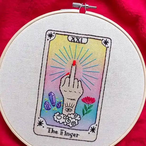 An embroidered tarot card in a hoop, depicting a hand with a middle finger raised, surrounded by crystals and a flower…