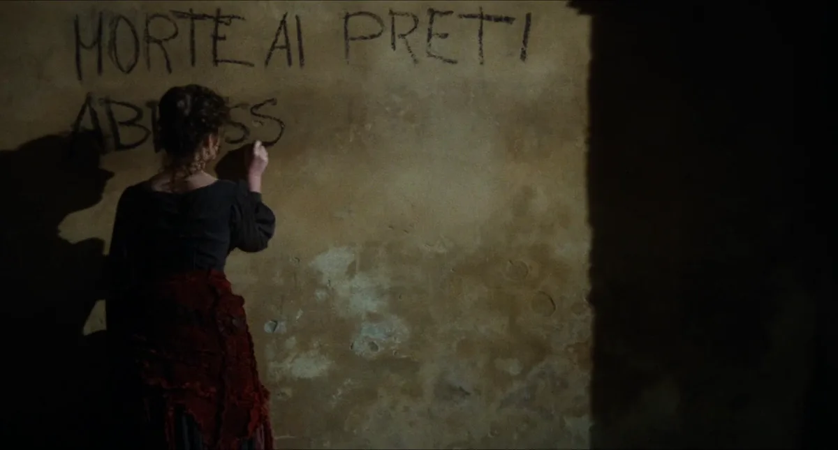 A woman in period clothing is seen from behind, writing graffiti on a rough, dimly lit wall. The text she writes reads…