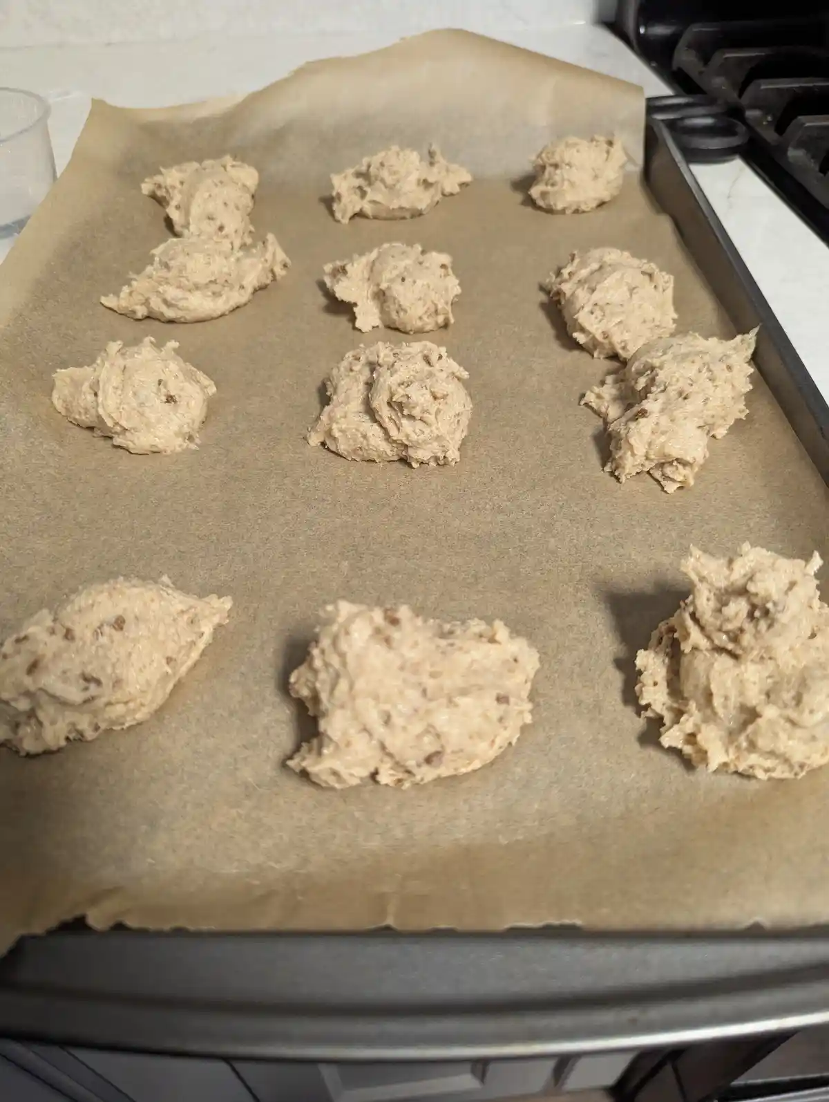 A baking sheet lined with parchment paper holds uneven, lumpy dollops of cookie dough arranged in rows. The dough is light…