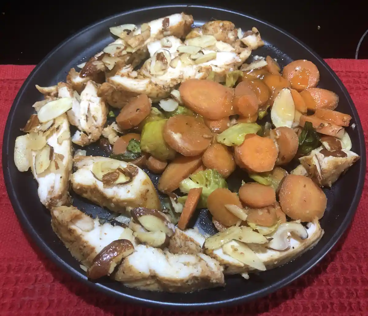 A black plate holds a meal of sliced chicken breast, sliced carrots, and Brussels sprouts, all coated in a glossy sauce and…