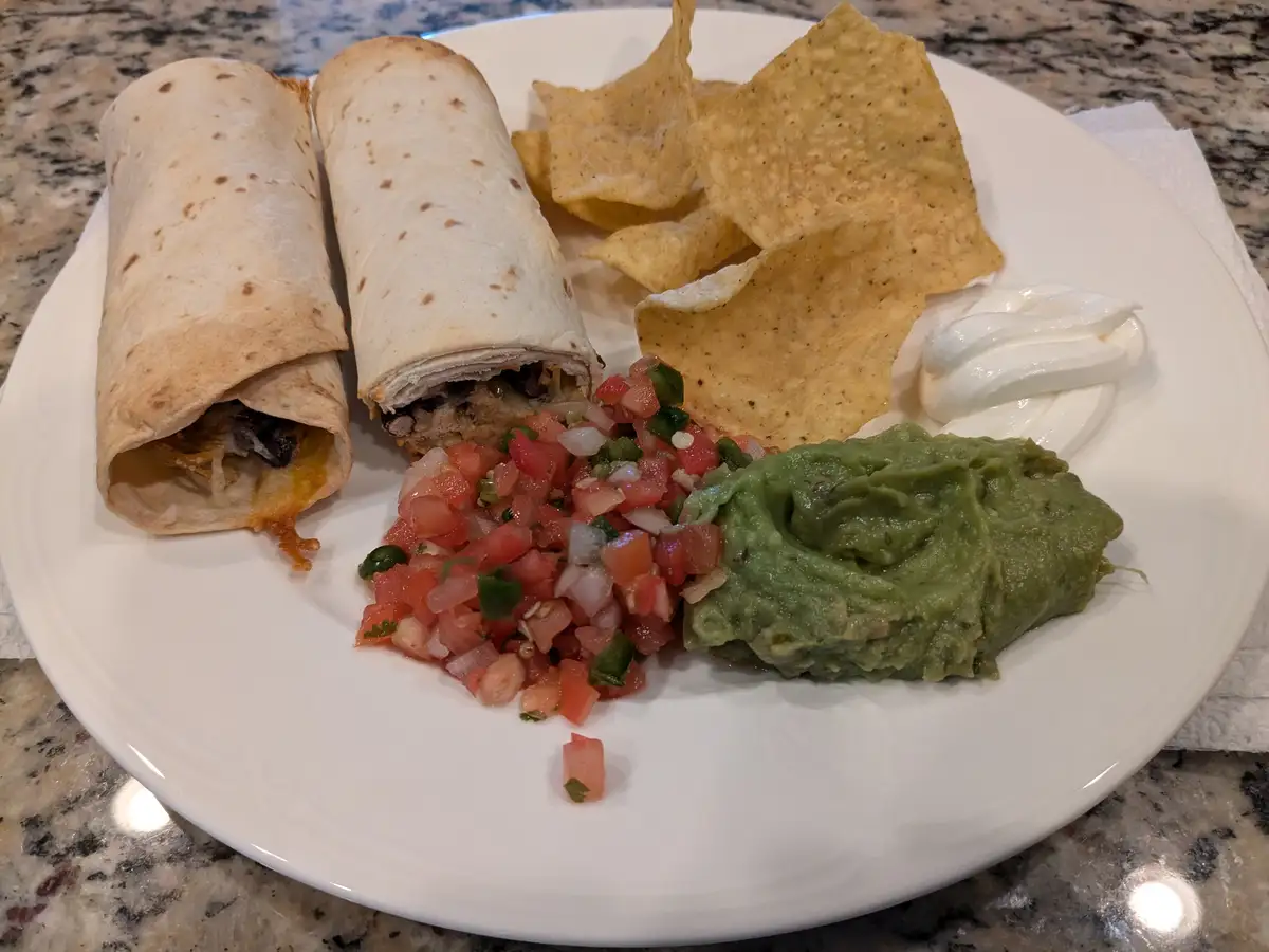 A plate of food featuring two burritos, tortilla chips, guacamole, salsa, and sour cream on a white plate. The overlaid text…