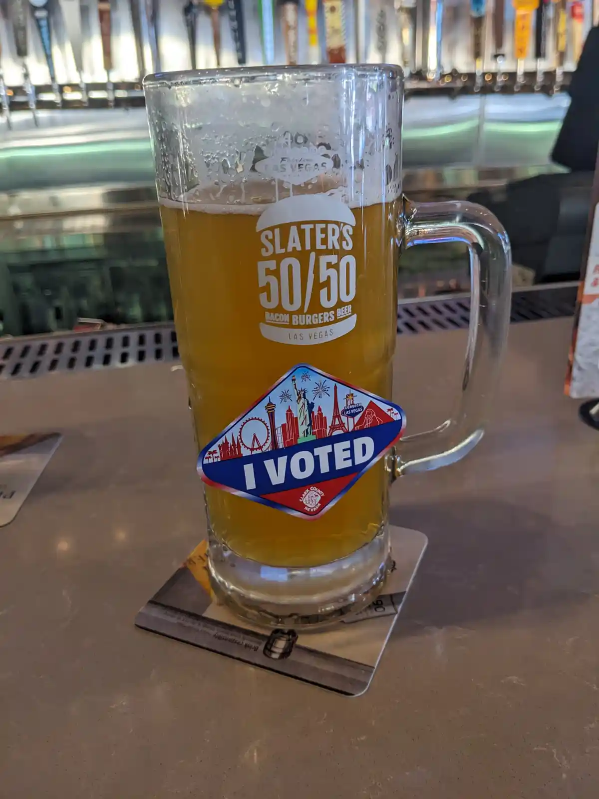 A tall glass mug of beer, branded with 'SLATER'S 50/50 BACON BURGERS BEER LAS VEGAS', sits on a bar counter with an 'I…