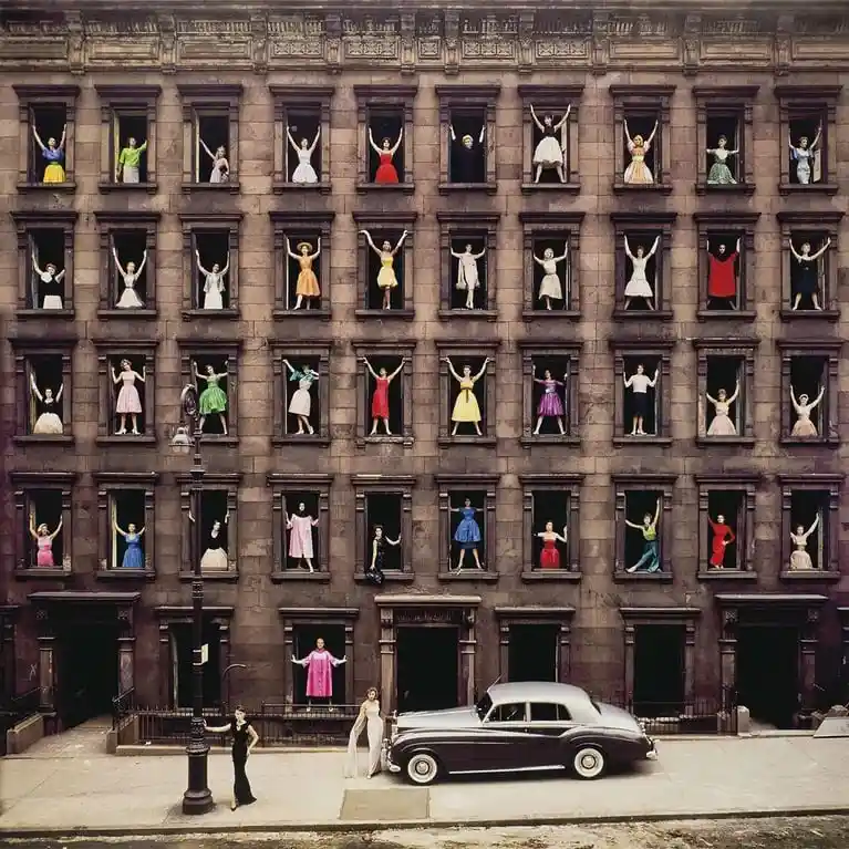 A vintage photograph shows a multi-story brownstone building with numerous people in colorful dresses and suits posing in…