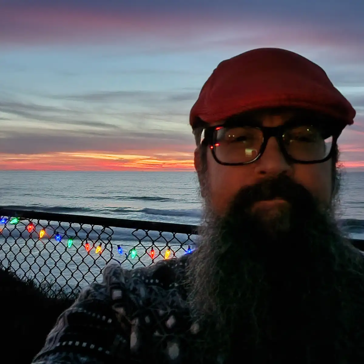 A man with a long beard, glasses, and a red cap takes a selfie at dusk overlooking the ocean, with a chain-link fence…