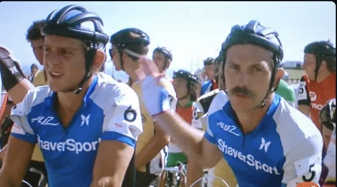 Two male cyclists in blue and white 'ShaverSport' jerseys with helmets are riding in a race; one cyclist on the right with a…