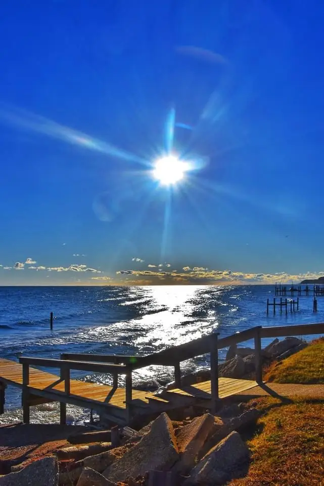 A bright, sun-drenched coastal scene with a wooden pier leading to the ocean under a clear blue sky with the sun shining…