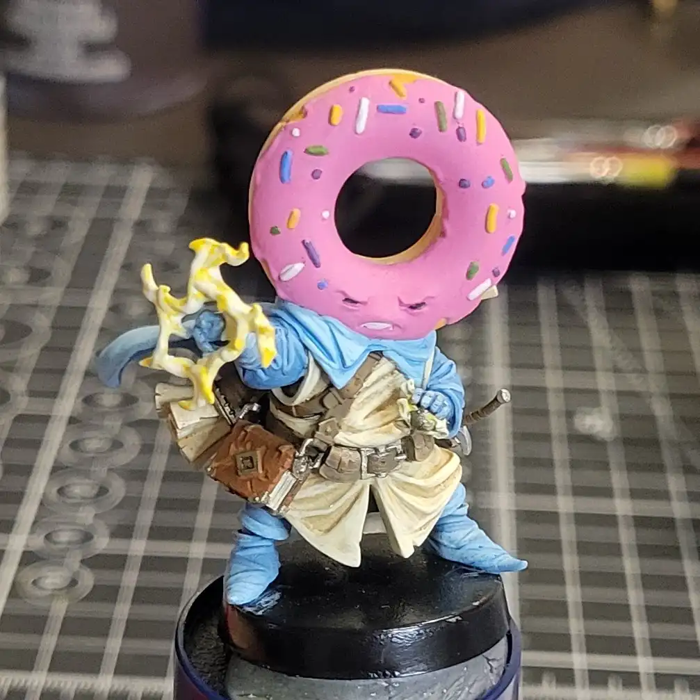 A painted miniature figurine of a fantasy character, dressed in blue and beige armor with a sword and glowing yellow weapon…