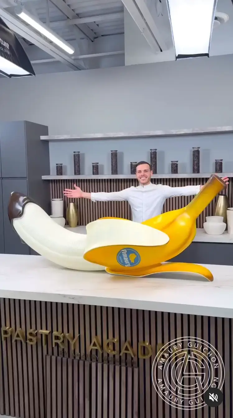 A smiling man in a white chef's coat stands behind a counter with arms outstretched, presenting a giant, realistic-looking…