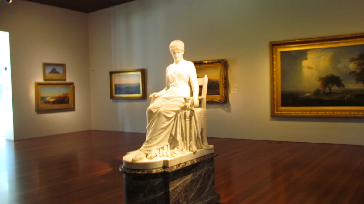 A white marble statue of a seated woman in classical robes is displayed on a dark marble pedestal in an art museum gallery…