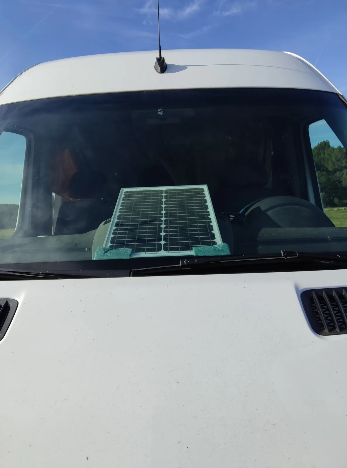 A small solar panel is taped to the inside of the windshield of a white van, positioned to face the sun under a clear blue…