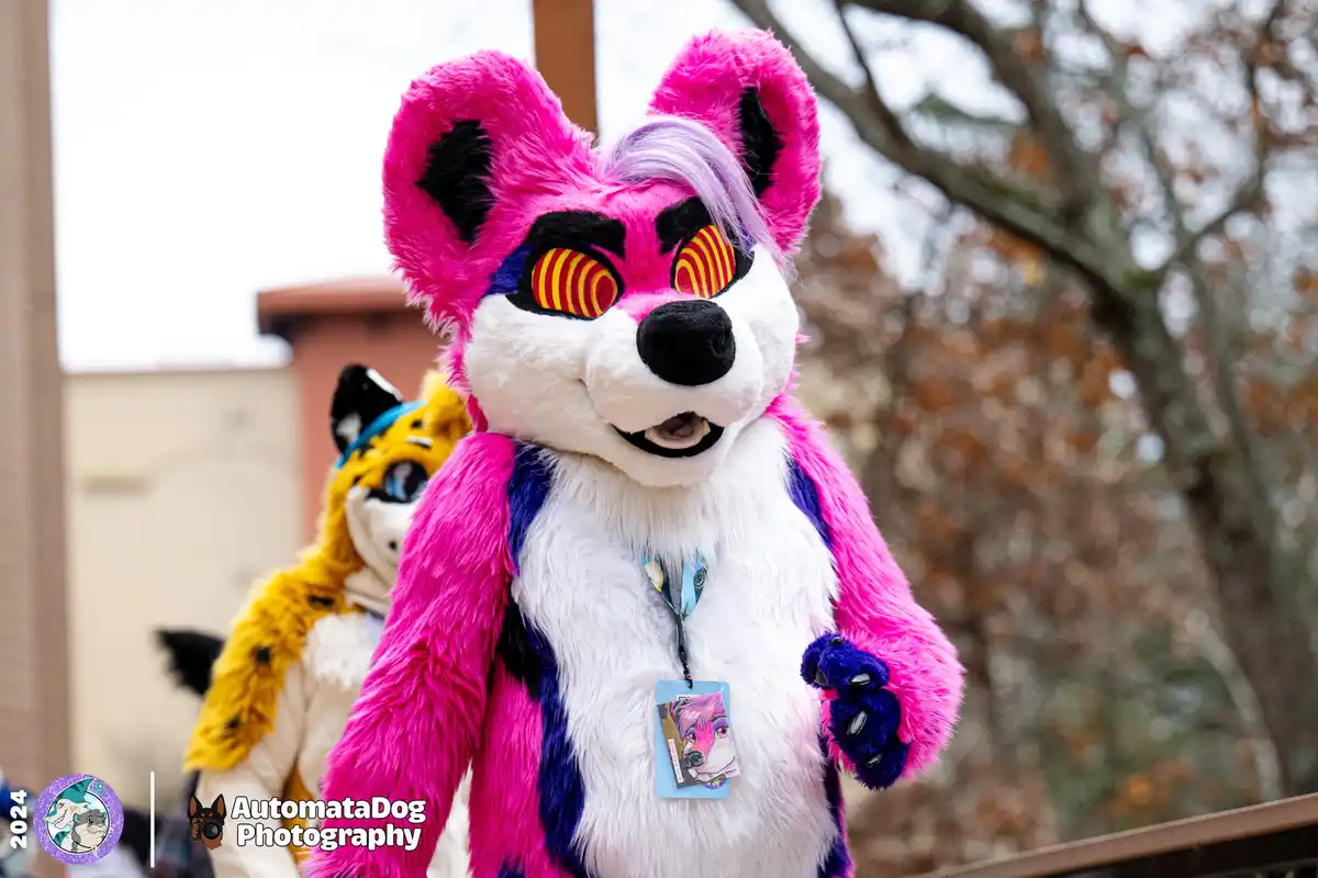 A person in a vibrant pink and white fursuit with striking red and yellow striped eyes and a purple streak in their hair…