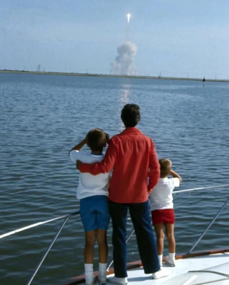 Three people, including two children, stand on a boat watching a rocket launch in the distance. The rocket is ascending with…