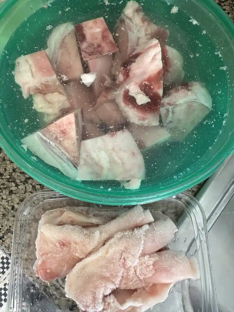 A green bowl filled with water and partially thawed, icy fish pieces sits above a clear plastic container holding more…