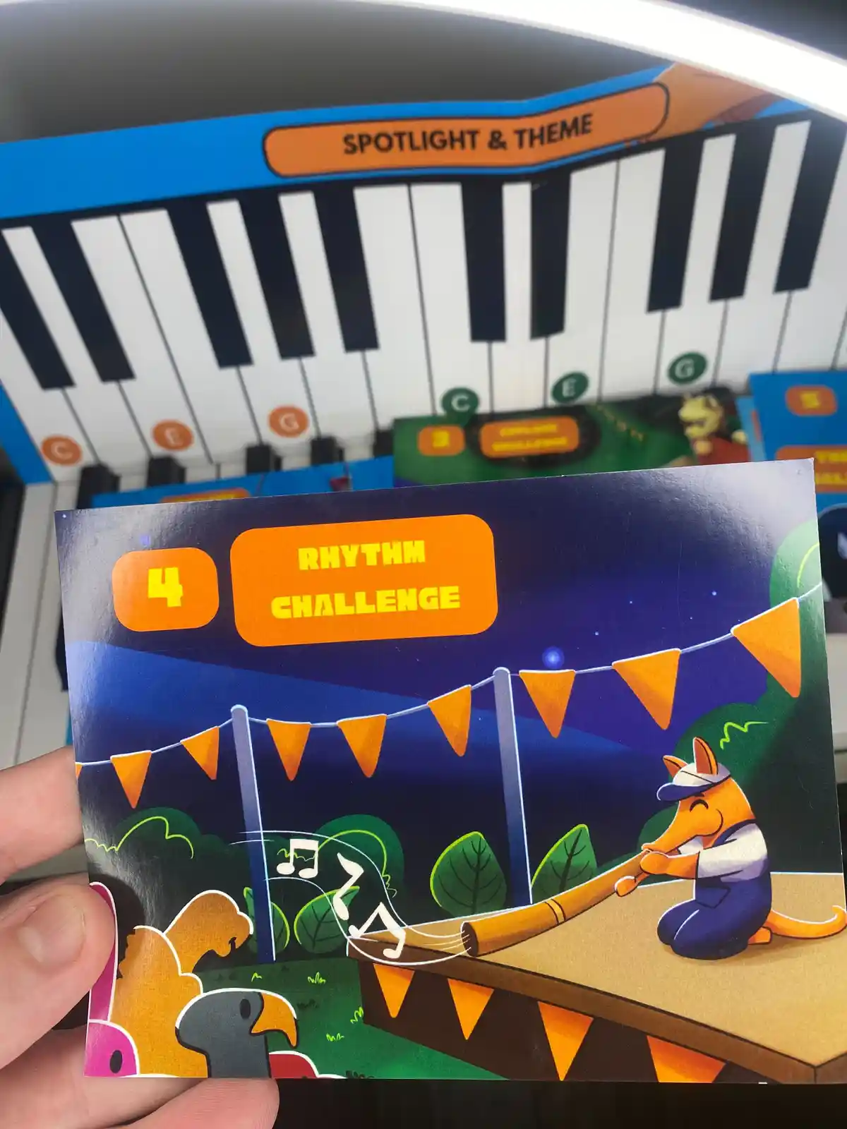 A hand holds a game card labeled '4 RHYTHM CHALLENGE' featuring a cartoon fox playing a didgeridoo under string lights at…