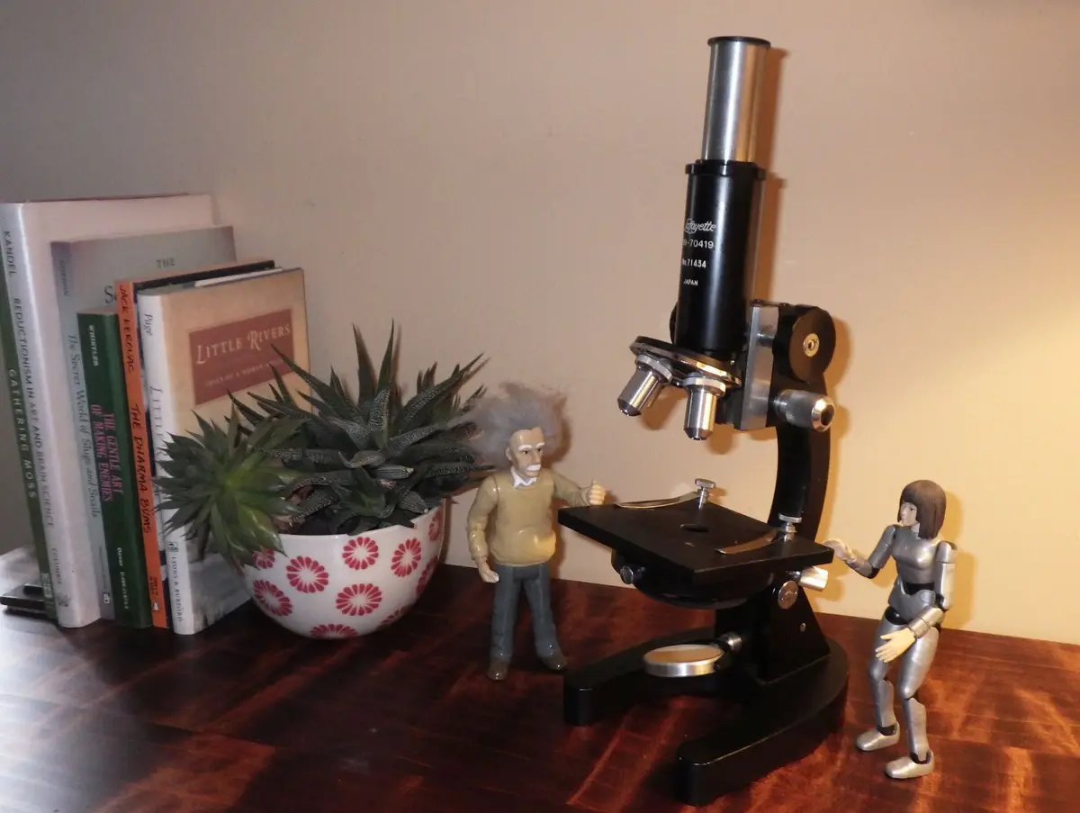 A photo shows a toy figurine of Albert Einstein gesturing toward a real microscope, while a silver humanoid robot figurine…