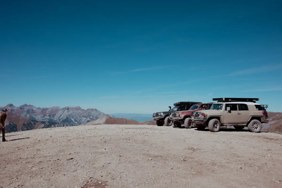 Three rugged off-road vehicles, including a beige Toyota FJ Cruiser, are parked on a dusty mountain ridge under a clear blue…