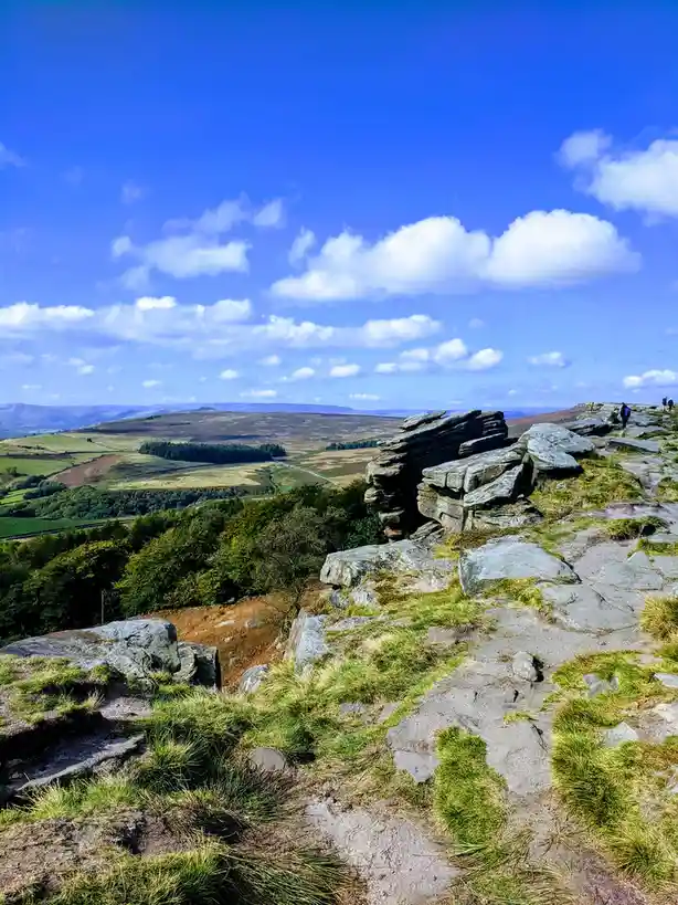 A scenic photo of a rocky hilltop with green grass and a vast landscape under a bright blue sky with fluffy white clouds…