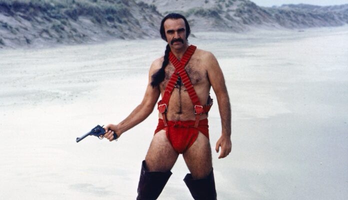 A man with a mustache and long dark hair in a ponytail stands in a desert-like landscape, wearing red suspenders and red…