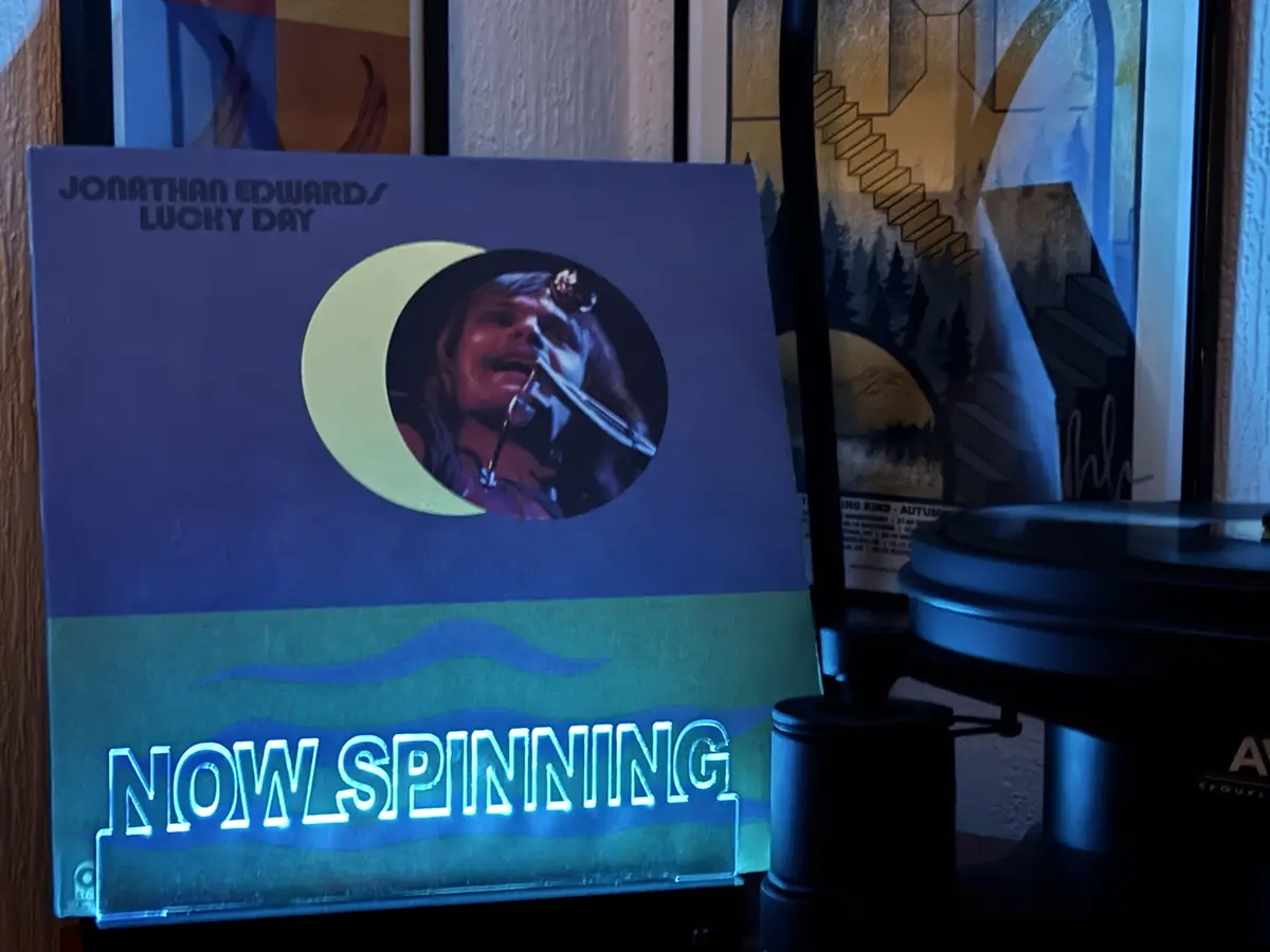A vinyl record album cover for 'Lucky Day' by Jonathan Edwards is displayed next to a turntable. The cover features a…
