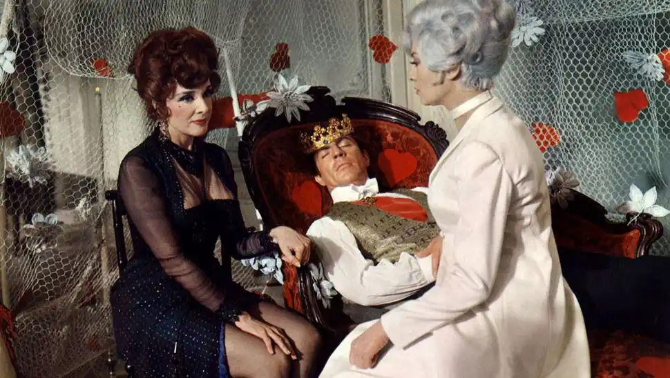 A still from a vintage film shows a man wearing a crown, reclining in a red upholstered chair with heart patterns, appearing…