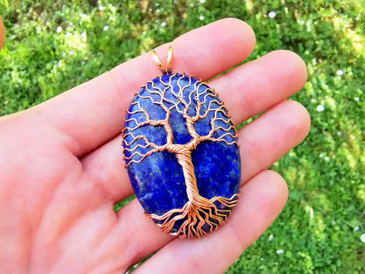 A hand holds a blue lapis lazuli pendant with a copper wire tree of life design against a green grassy background. The…