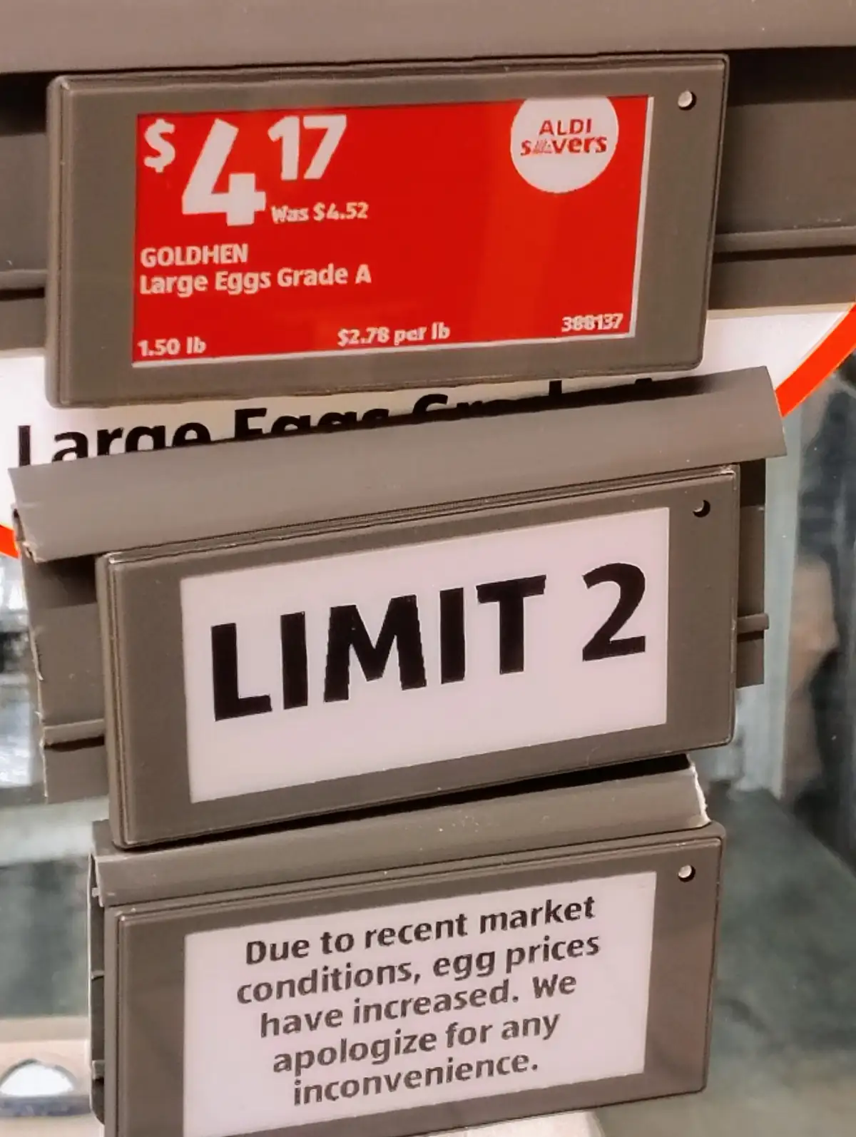 A close-up of three stacked Aldi price signs for eggs. The top red sign shows a price of $4.17 for 1.50 lb of Goldhen Large…