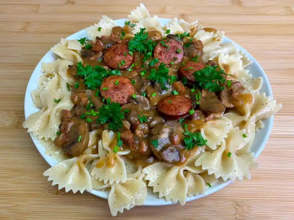 A white plate holds bow-tie pasta topped with a brown gravy containing sliced sausage, mushrooms, and garnished with chopped…