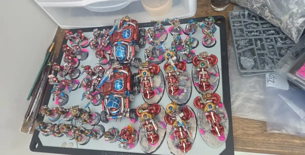 An overhead photo shows a large collection of intricately painted miniature figurines, mostly in red, blue, and gold…