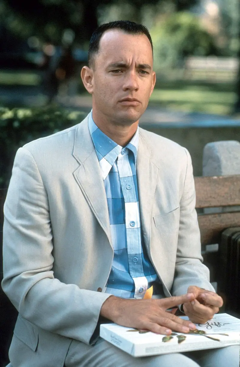 tom hanks sits on a park bench wearing a light gray suit over a blue and white checkered shirt, looking displeased while…