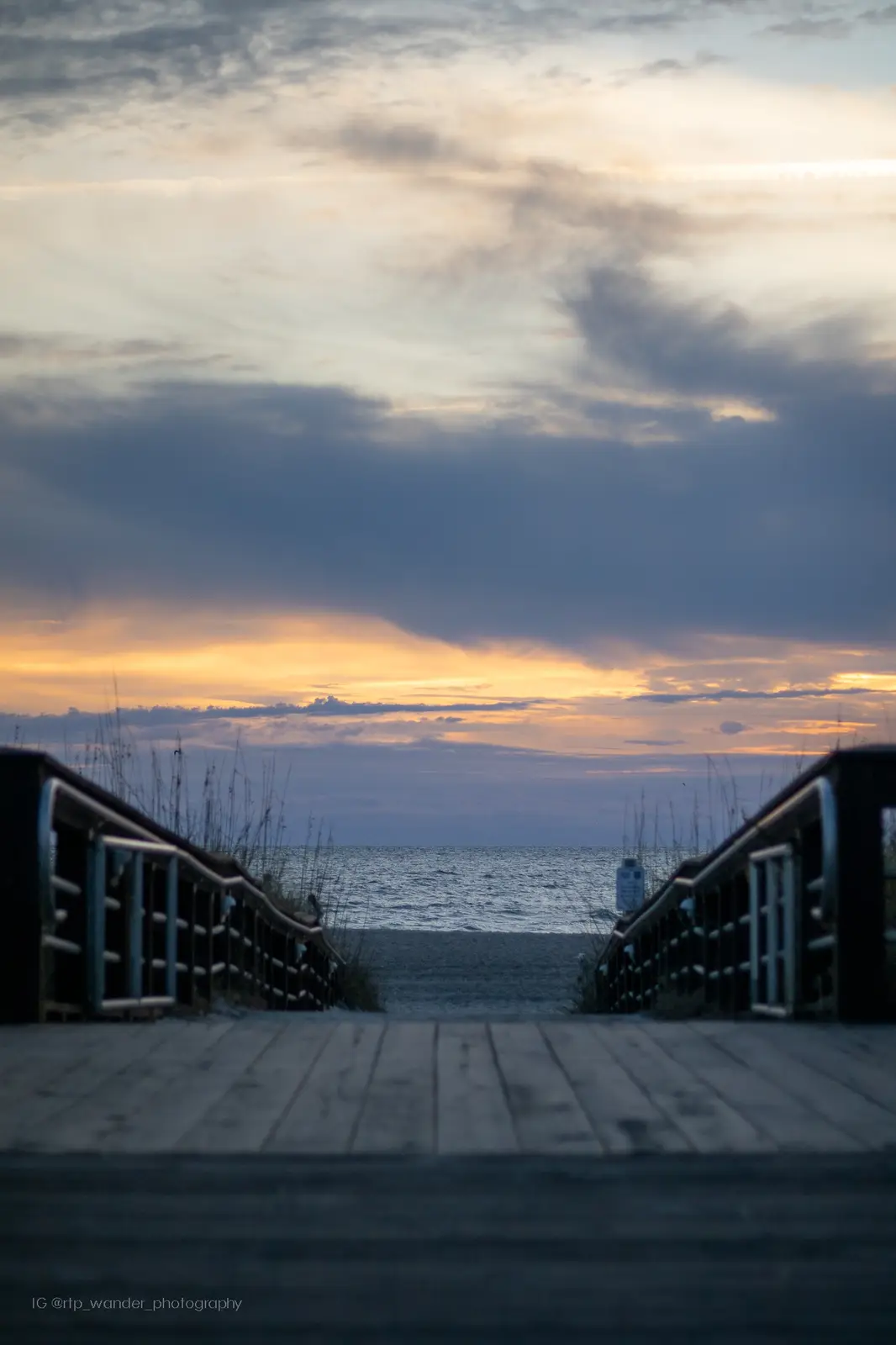 A serene photograph shows a wooden boardwalk leading down to a beach at sunset, with dramatic clouds and orange light on the…