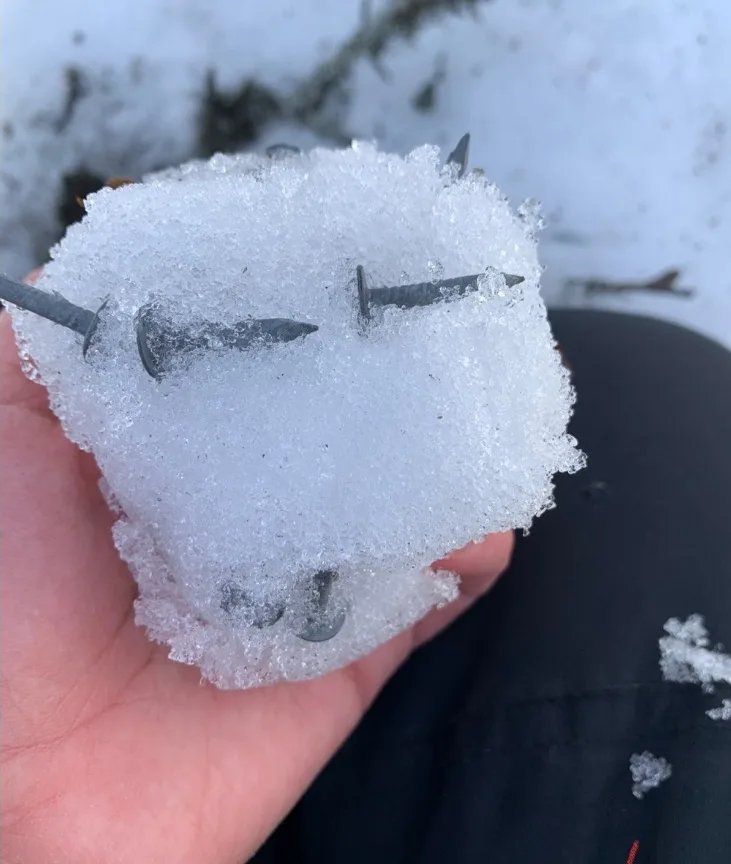 A hand holds a snowball shaped like a face, with several metal nails embedded to form eyes, a nose, and a mouth. The…