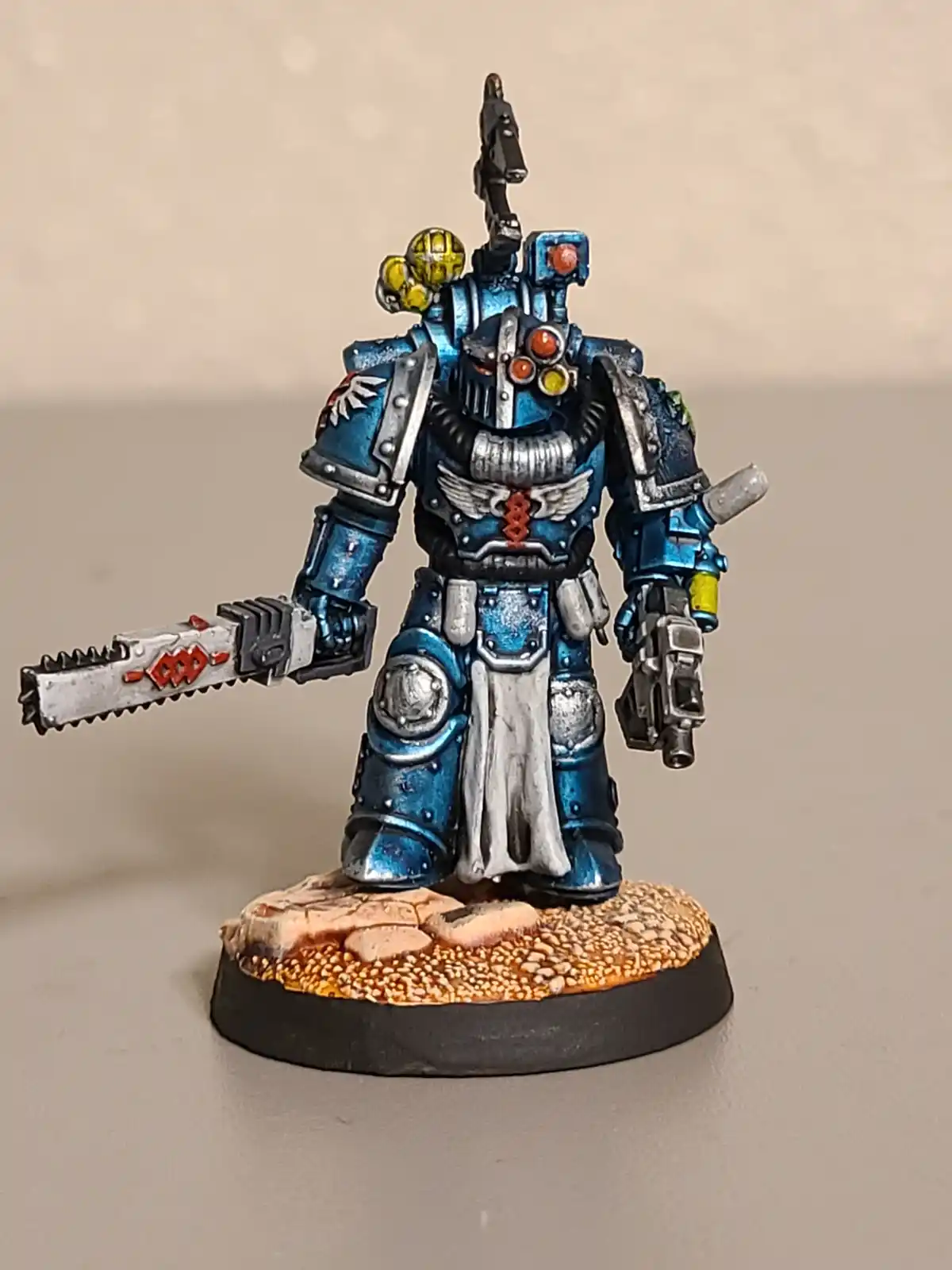 A painted miniature of a blue-armored Space Marine from Warhammer 40,000 stands on a rocky base, holding a chainsword in one…