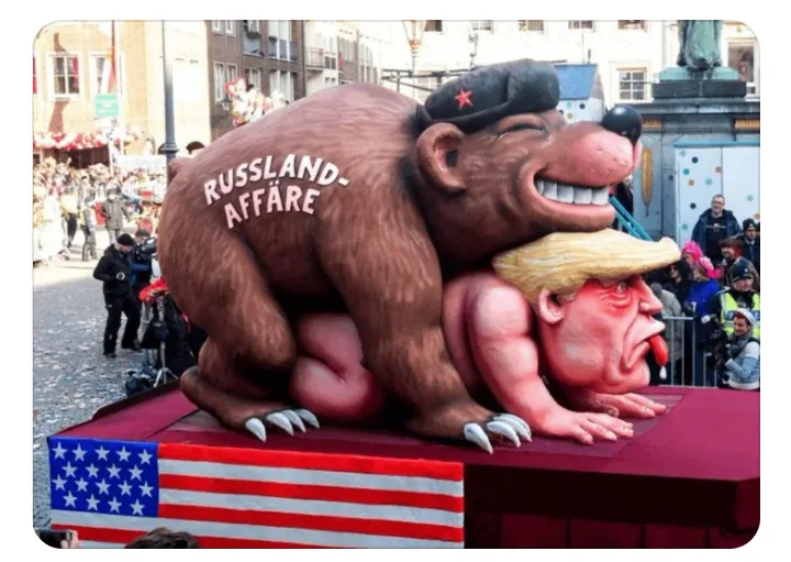 A large parade float depicts a cartoonish brown bear wearing a black military cap with a red star, mounted on top of a…