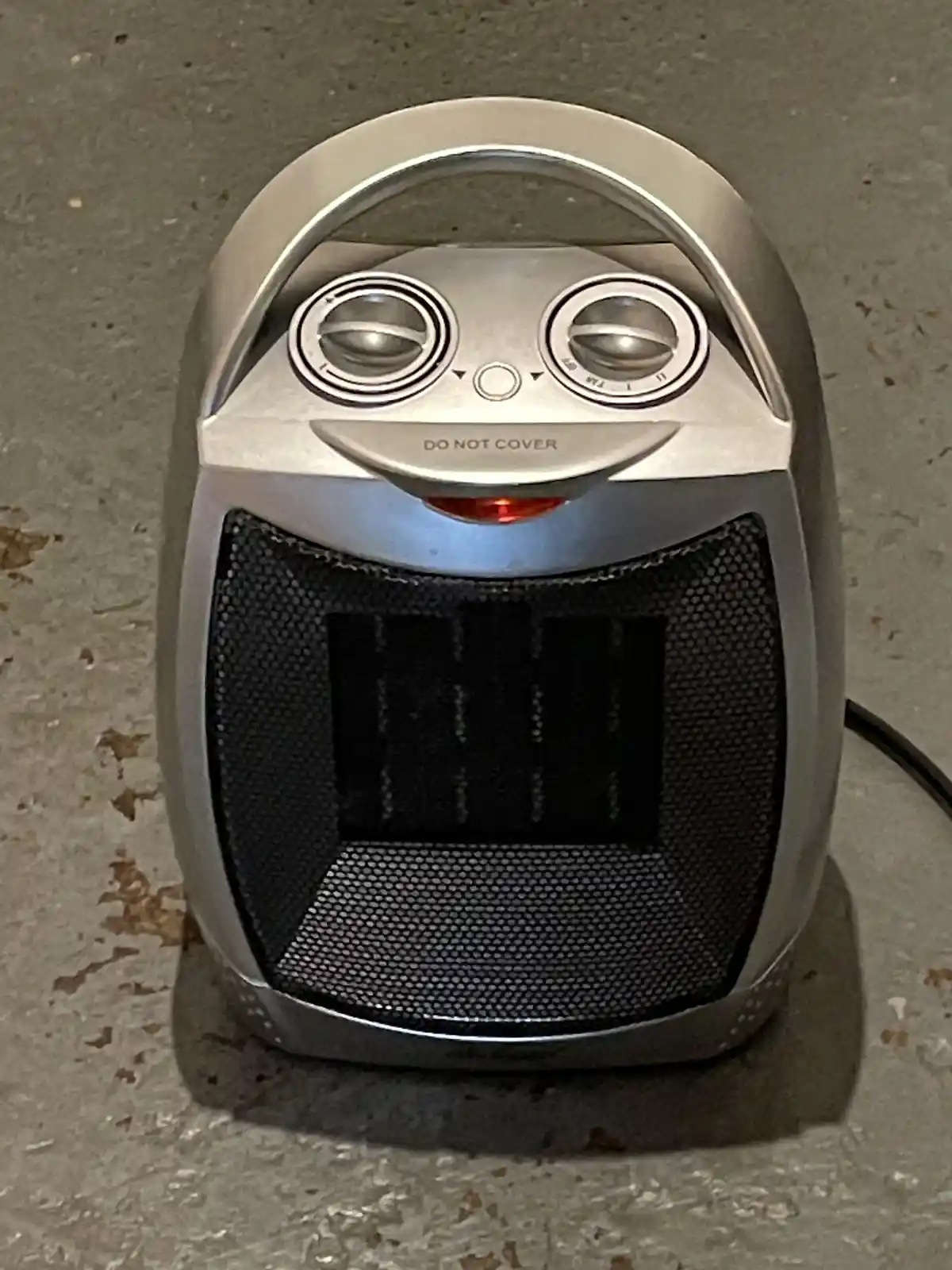 A silver space heater with two dials and a red power light sits on a concrete floor, its control panel humorously edited to…