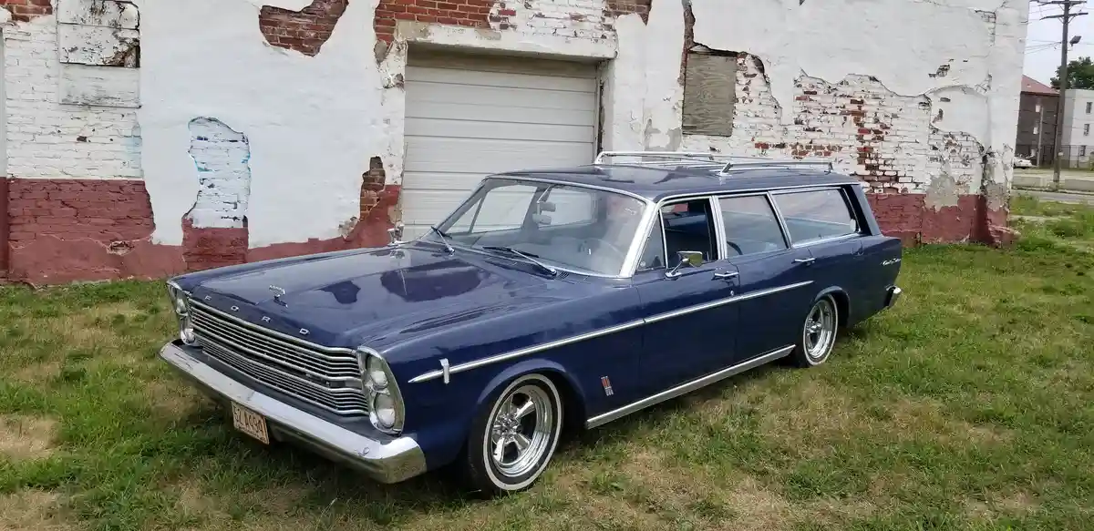 A well-maintained blue 1960s Ford station wagon with chrome trim and custom wheels is parked on grass in front of a…