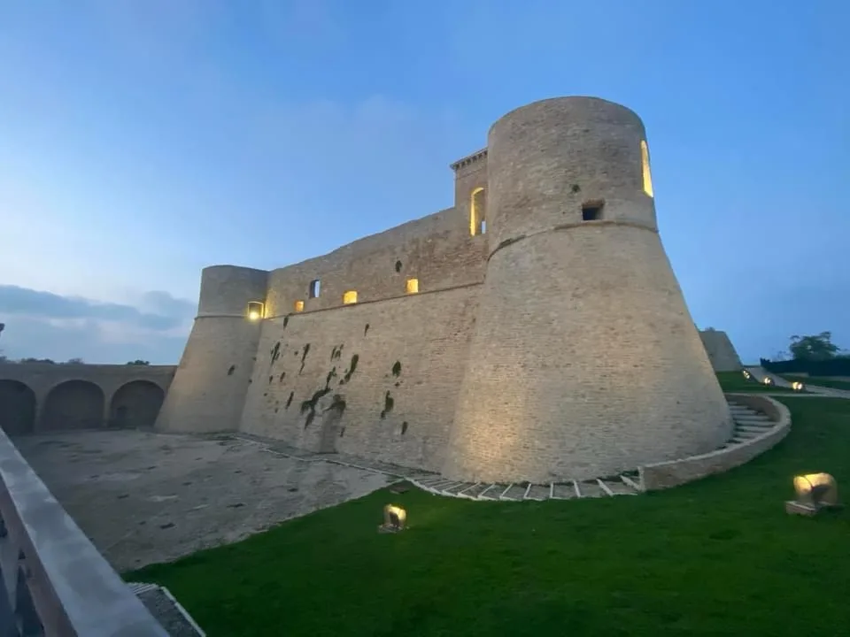 A large, ancient-looking stone castle with cylindrical towers and illuminated windows stands at dusk, surrounded by green…