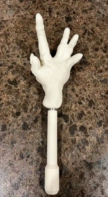 A white, plastic-looking hand attached to a long, cylindrical handle rests on a speckled brown countertop. The hand is posed…