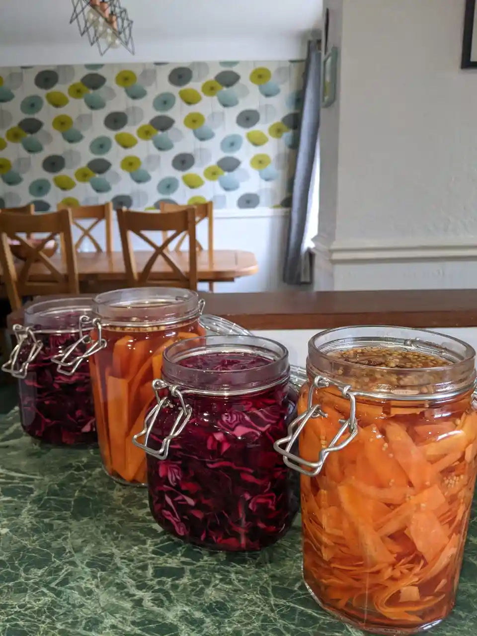 Four glass jars with clamp lids sit on a green marble countertop, filled with pickled vegetables. Two jars contain vibrant…