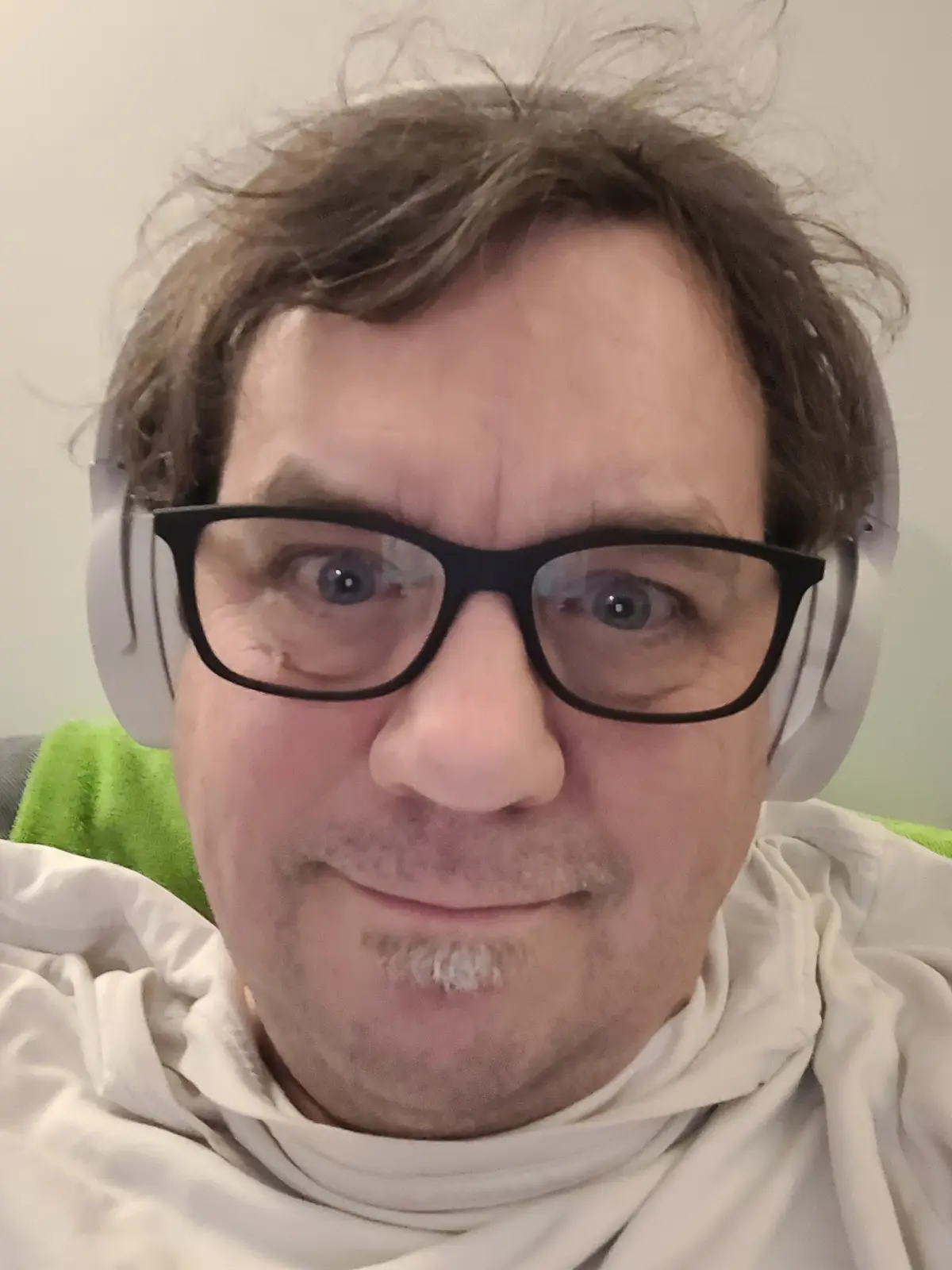 A close-up selfie of a man with messy brown hair, wearing black-rimmed glasses and white over-ear headphones, looking…