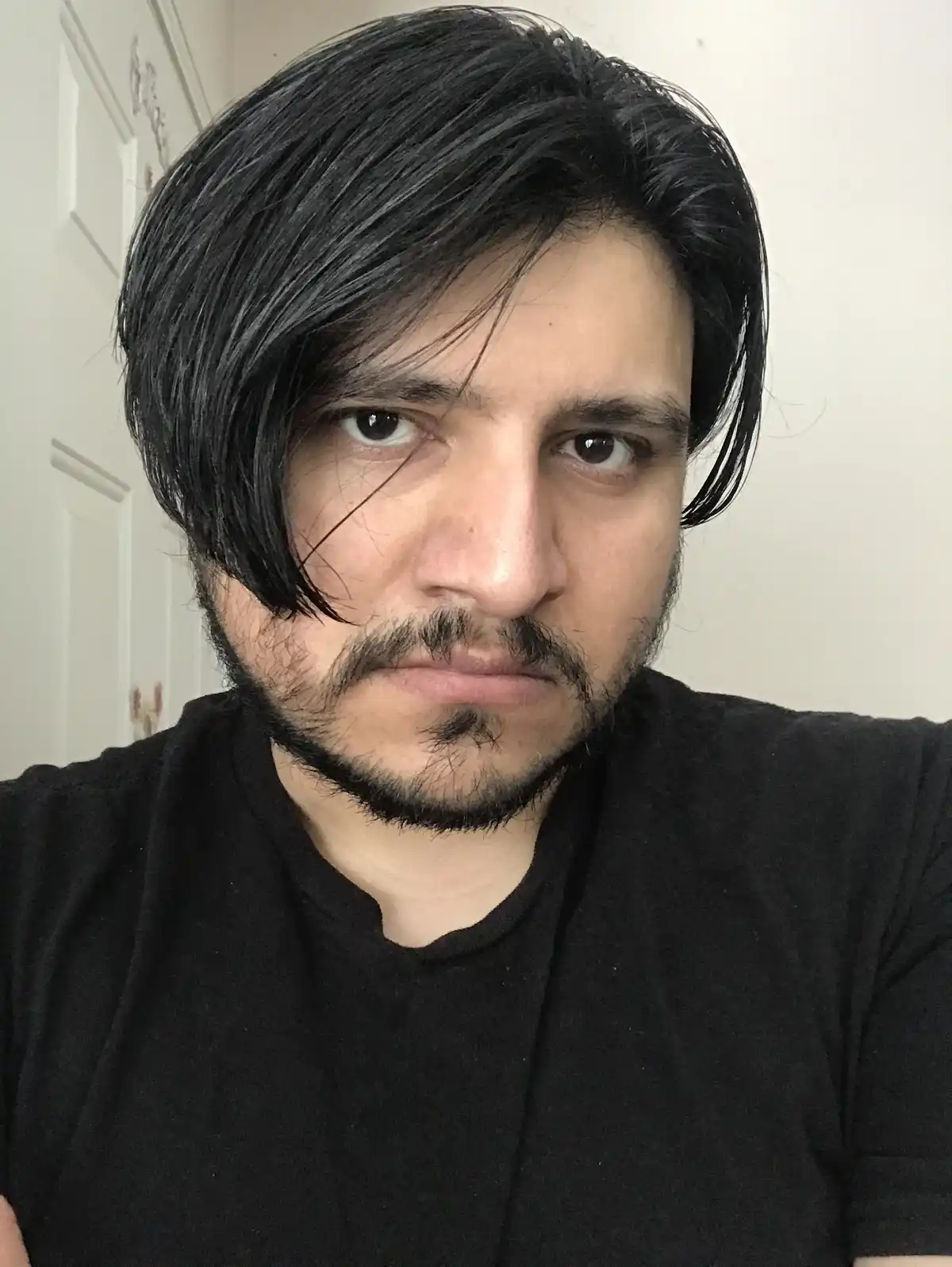 A man with dark, styled hair and a goatee stares intensely at the camera while wearing a black t-shirt. The overlaid text…