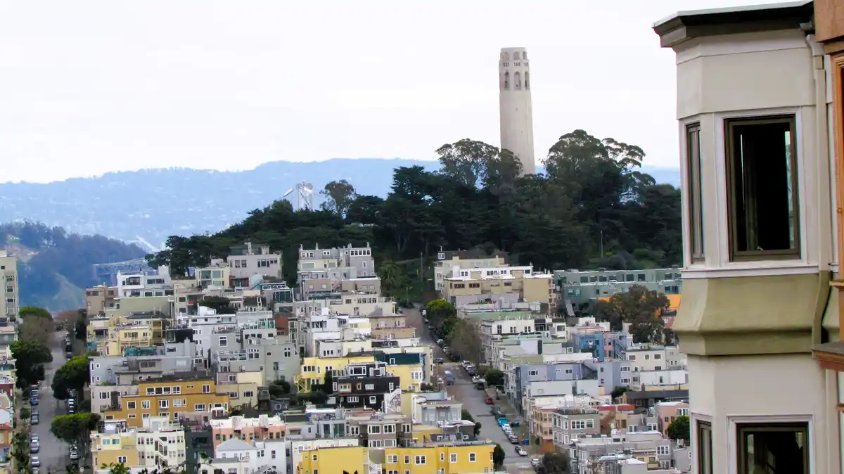 A scenic view of San Francisco showing Coit Tower on a hill, colorful houses on steep streets, and the Bay Bridge in the…
