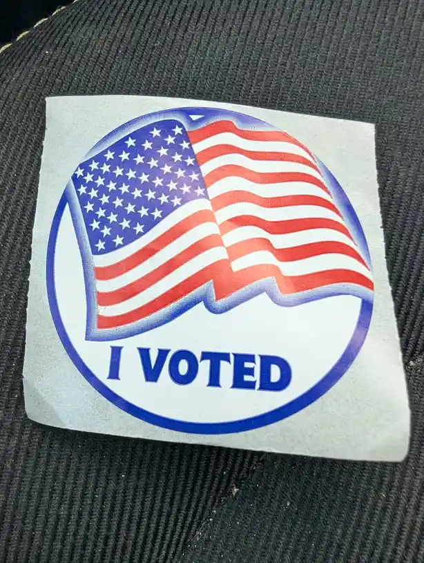 A close-up photo shows a square sticker with a circular design featuring a waving American flag and the text 'I VOTED' in…