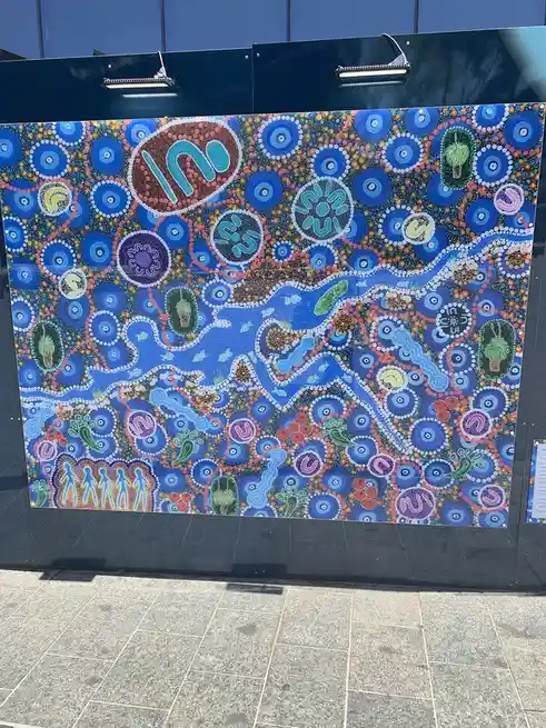 A vibrant, large-scale Aboriginal dot painting featuring blue rivers, circular patterns, and stylized figures, displayed…