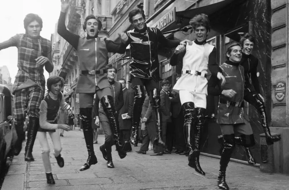 A black-and-white photo shows a group of men in flamboyant 1960s futuristic costumes, including shiny boots and metallic…
