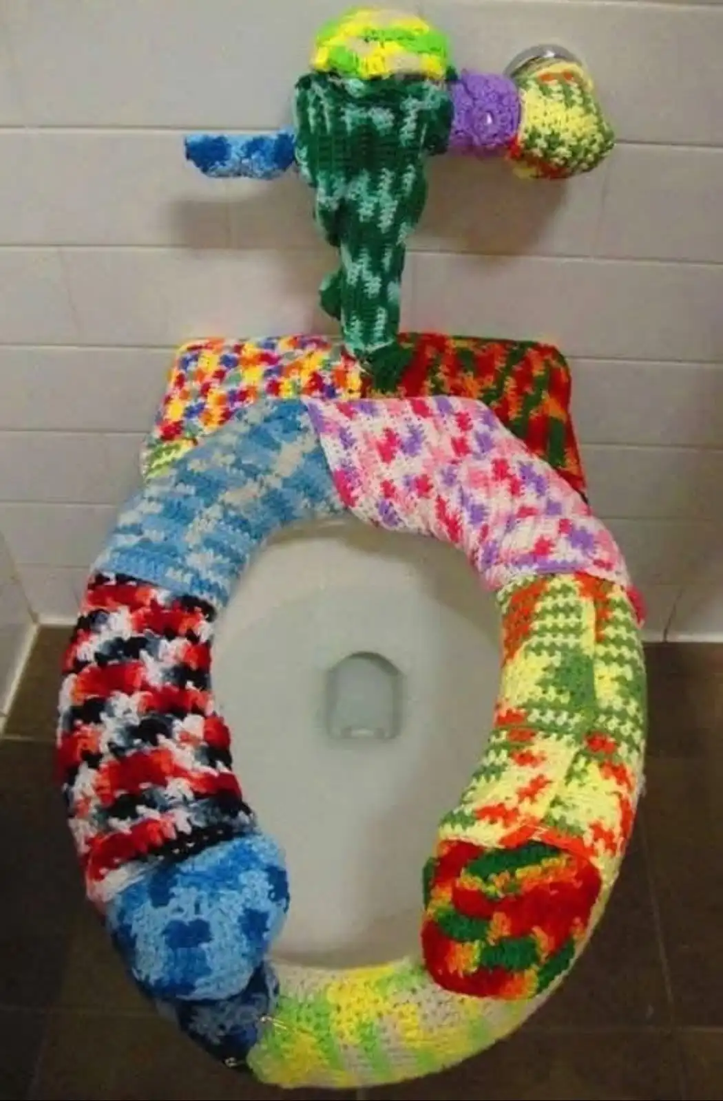 A toilet seat is entirely covered in a patchwork of colorful, hand-knitted fabric with various patterns and hues. Above the…