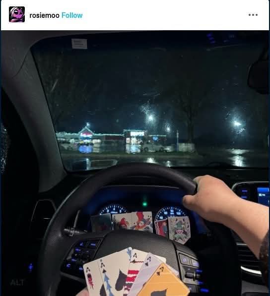 A person is driving at night in a car with rain on the windshield, holding four aces in their hand. The dashboard has more…