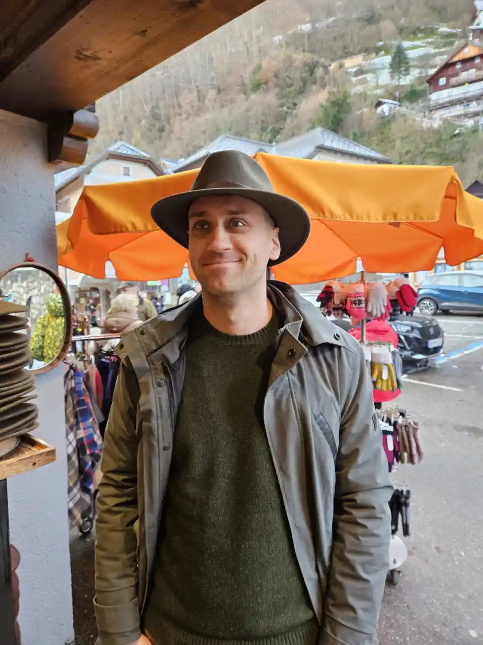 A man wearing a brown fedora, olive green sweater, and matching jacket stands in front of an outdoor market stall with…