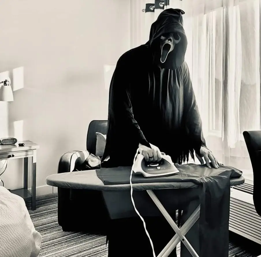 A person wearing a black hooded costume with a white screaming face mask is standing in a hotel room, holding an iron over a…